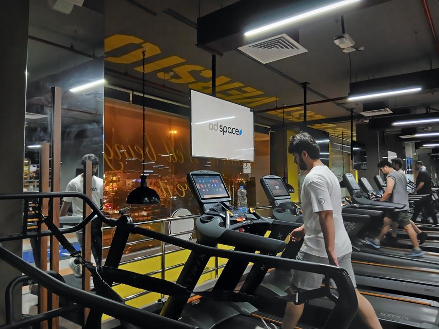 An Ad Space screen above the treadmills, with members running, in a men's gym in Bin Omran, Doha