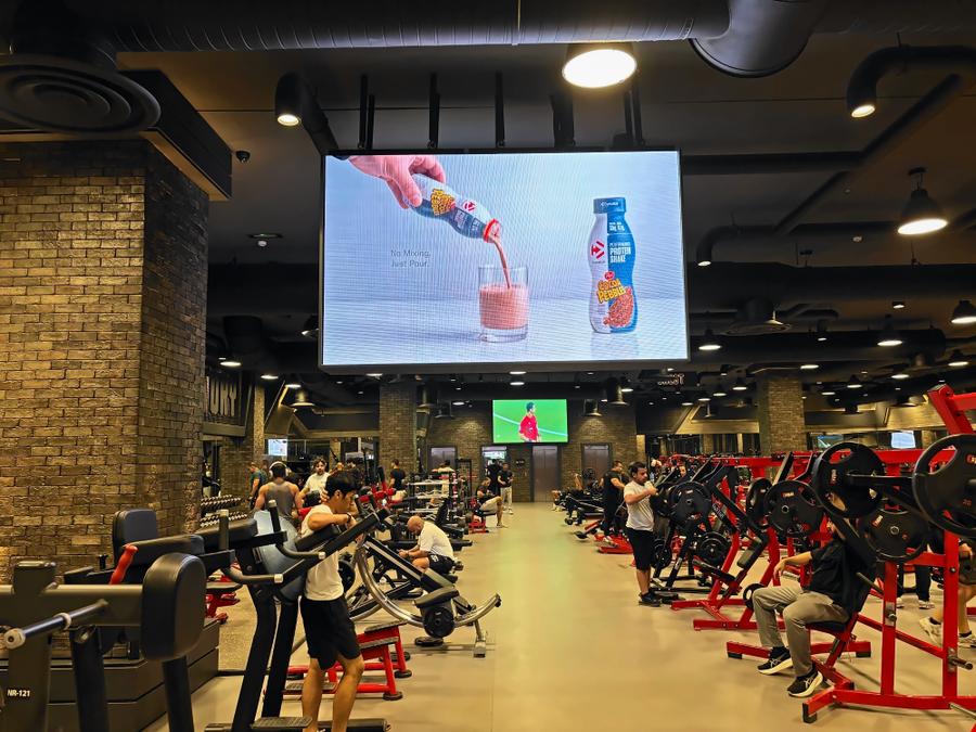 A drink ad on a giant screen over a busy training floor in a men's gym in Riggae, Kuwait
