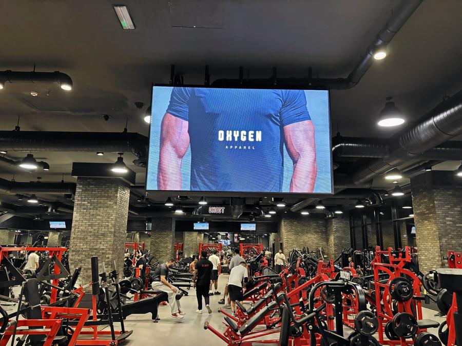 A giant screen playing a sportswear ad above the red machines of a busy men's gym in Sabah Al Salem, Kuwait