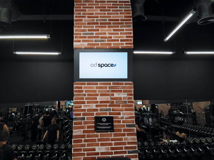 An Ad Space screen on a brick pillar above the cardio machines of a mixed gym in Duhail, Doha