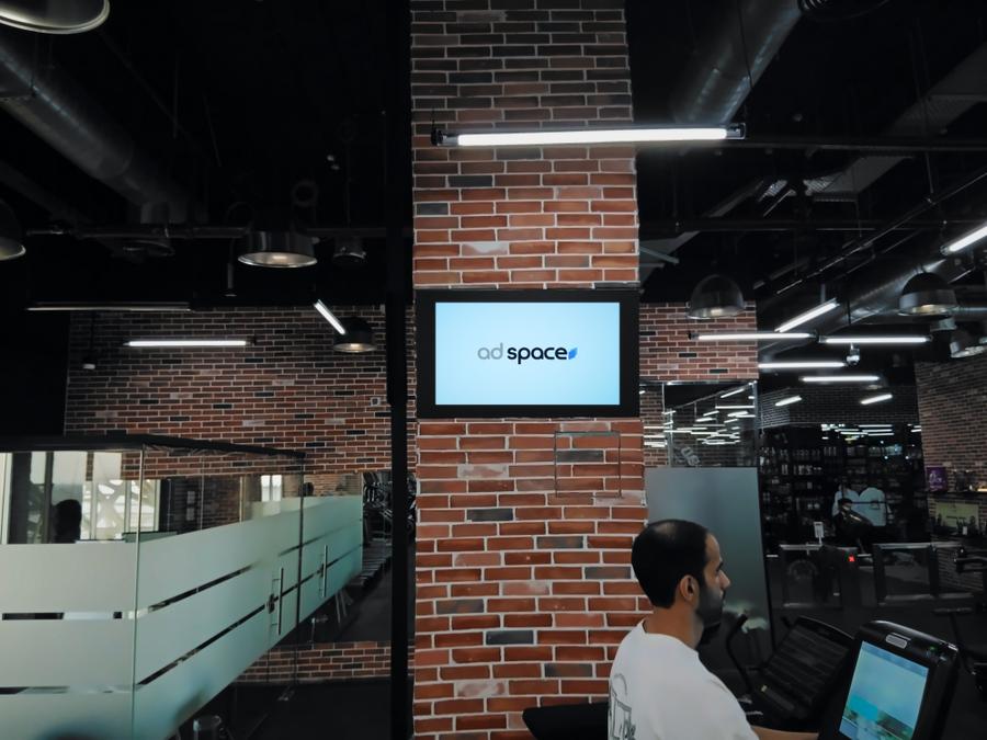 An Ad Space screen on a brick pillar above the treadmills of a mixed gym in Duhail, Doha
